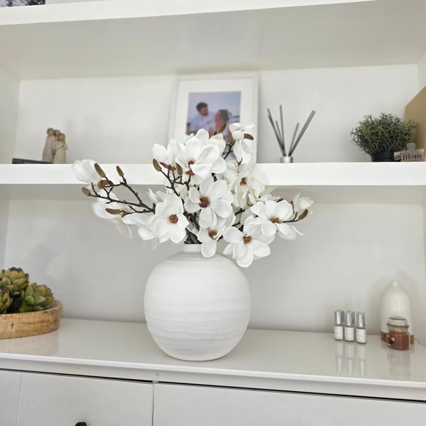 Faux White Magnolia Stems sat inside a white round ceramic vase sat on a white sideboard in a living room.
