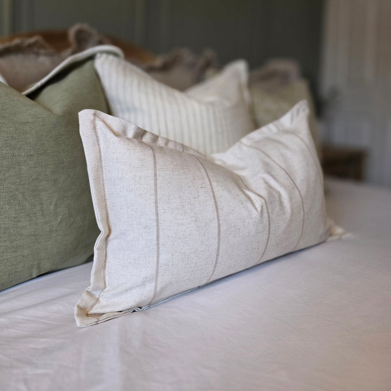 A rectangle cushion with a flanged edge. Cream, stone colour with a subtle vertical beige stripe repeating from left to right. Sat on a bed with white bedding
