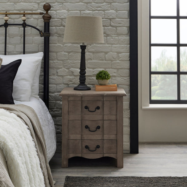 Cotswold 3 Drawer Wooden Bedside Table. Each drawer displays a black metal handle. Displaying a black base and cream shade table lamp on top. Sat at the side of a black metal frame bed.